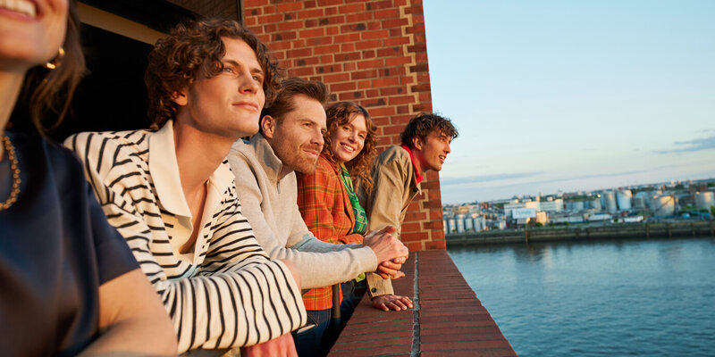 Eine Gruppe Menschen schaut vom Balkon eines Backsteingebäudes auf die Elbe