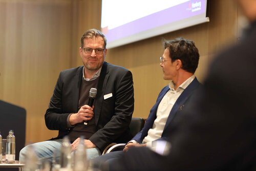 Zwei Männer diskutieren auf einer Veranstaltung, Mikrofon in der Hand. Impressionen der Veranstaltung "Exzellenz durch Kongresse" mit Podiumsdiskussion und Networking.